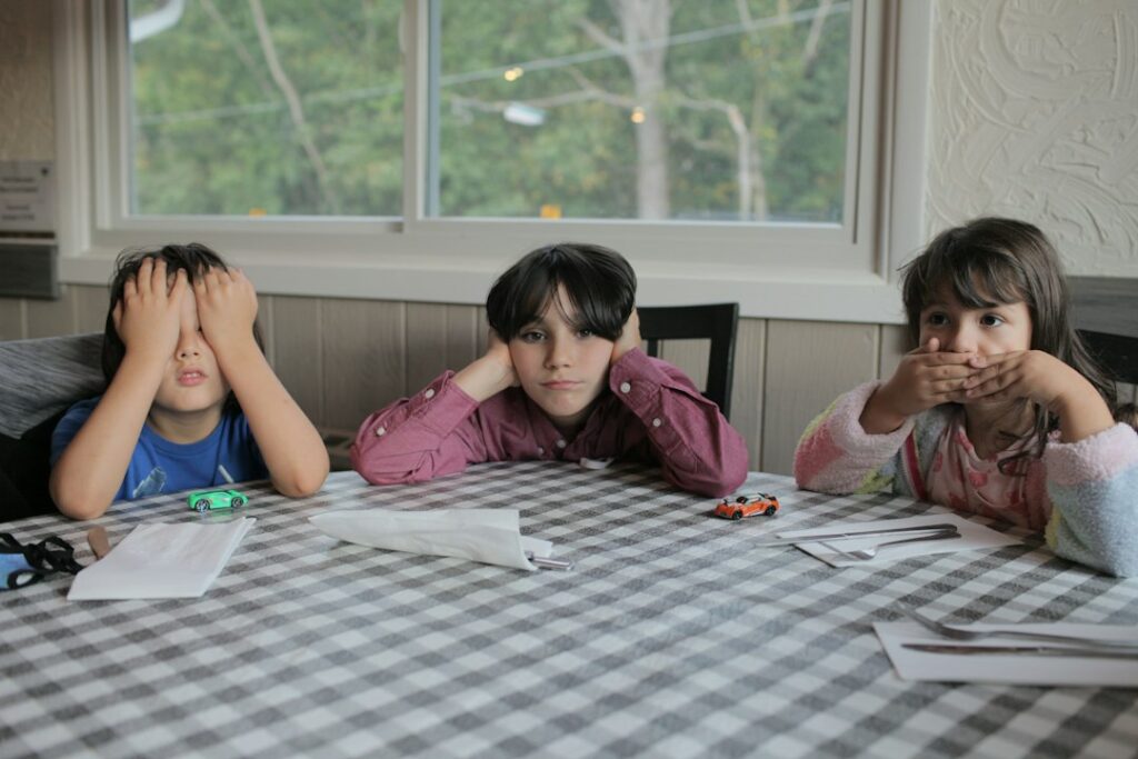 Two children read together at a kitchen table while one follows along with an adult reading aloud.