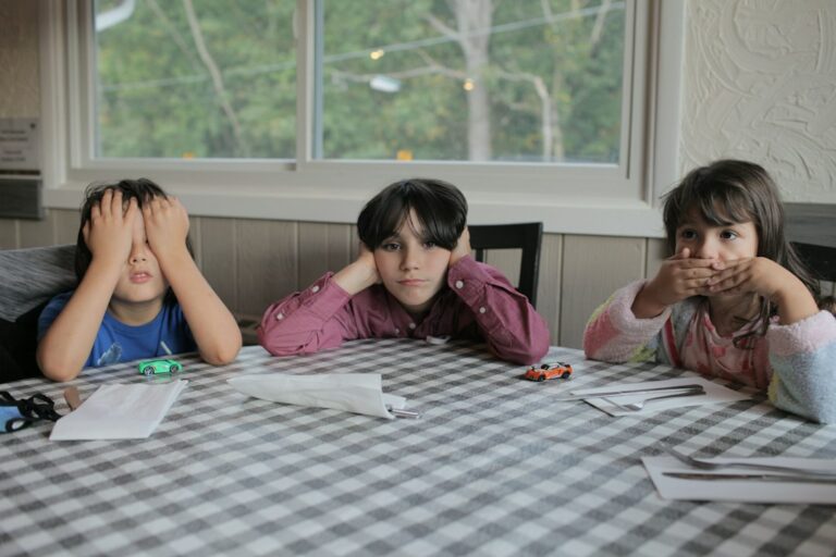 Two children read together at a kitchen table while one follows along with an adult reading aloud.